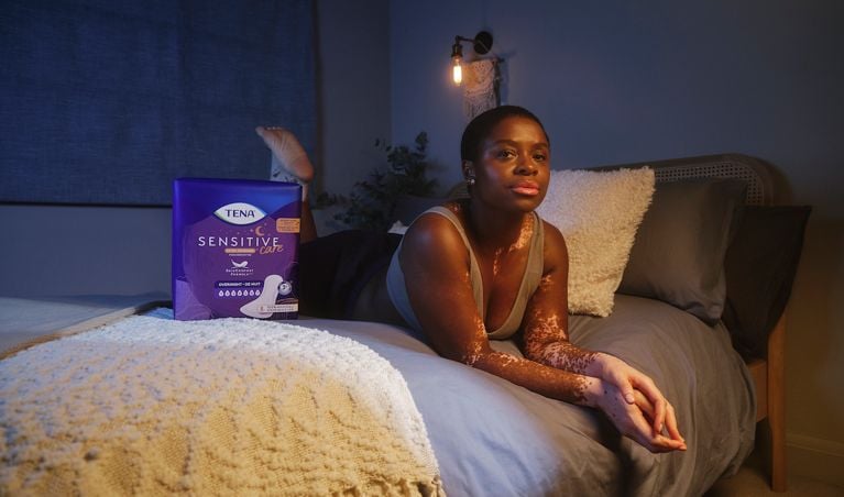 Woman lying on her stomach across a bed with a pack of TENA incontinence pads next to her.