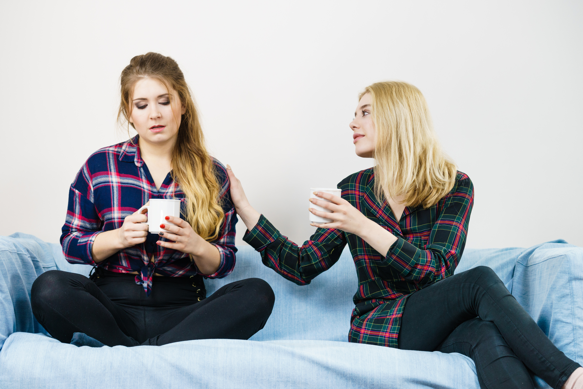 Two women sitting on a blue couch, both holding mugs. The woman on the left, with long brown hair, looks sad and is looking down. The woman on the right, with blonde hair, appears to be comforting her by placing a hand on her shoulder. Both are wearing plaid shirts.