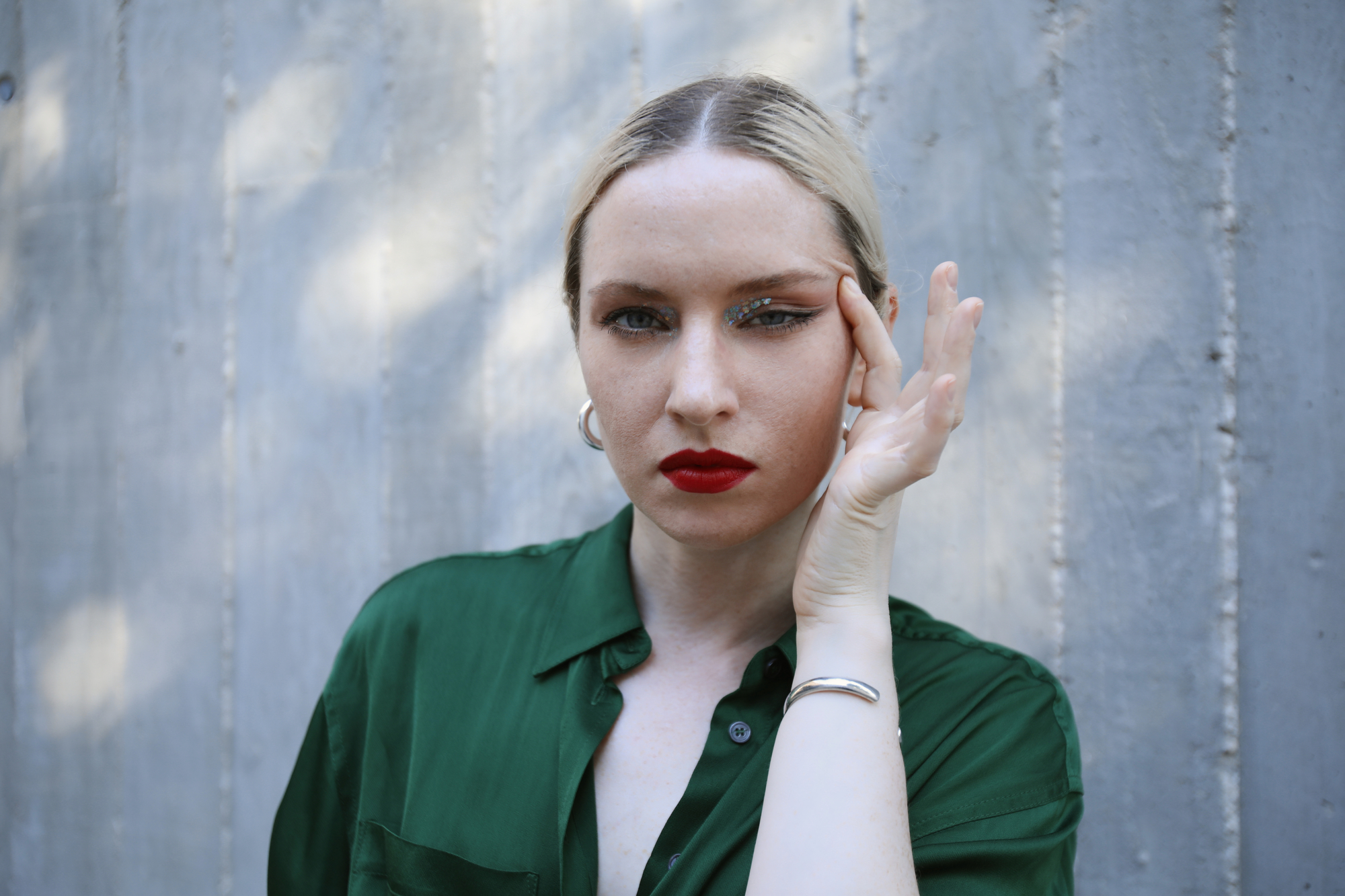 A person with blonde hair, pulled back, wearing a green blouse and hoop earrings, poses with a hand near their face. They have bold red lipstick and are in front of a gray, textured background.