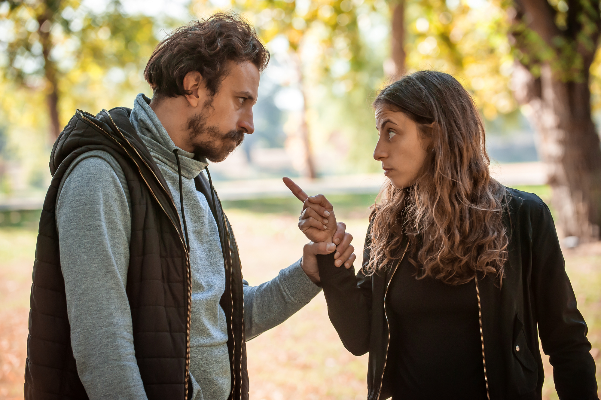 A man and a woman are outdoors in a park, engaged in a serious conversation. The woman, with long brown hair, is pointing her finger at the man while making an expressive face. The man, with short curly hair, is holding her wrist, looking at her intently.