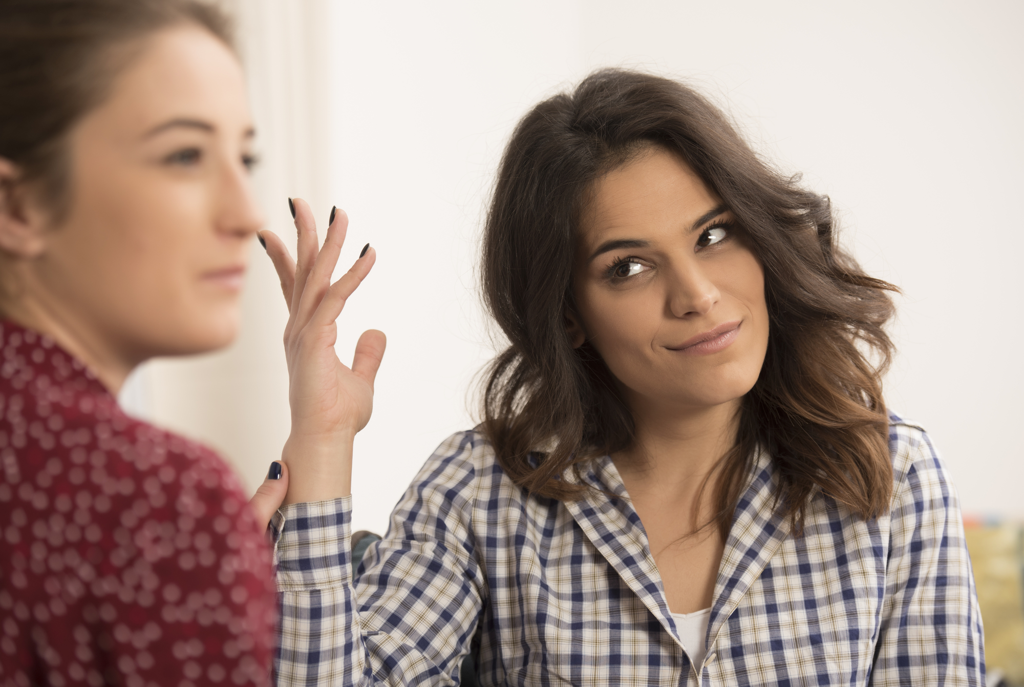 Two women are indoors having a conversation. One woman with brown hair and wearing a red patterned top looks away, while the other woman with wavy hair and wearing a checkered button-up shirt makes a gesture with her hand, appearing a bit skeptical or bemused.