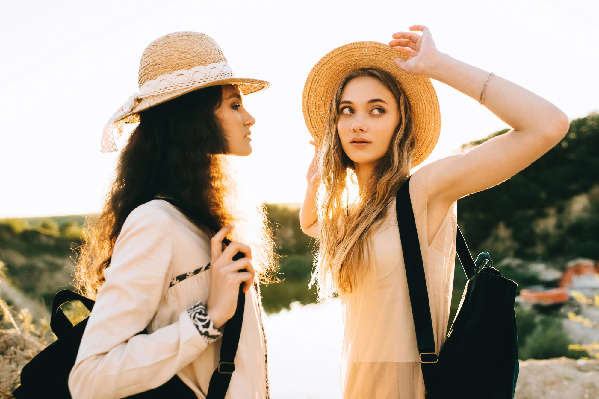 Two young women are standing outdoors, wearing straw hats and backpacks. They are dressed in light, summery clothing, with one adjusting her hat while looking to the side. The background is a scenic view with a bright, sunlit sky and natural surroundings.