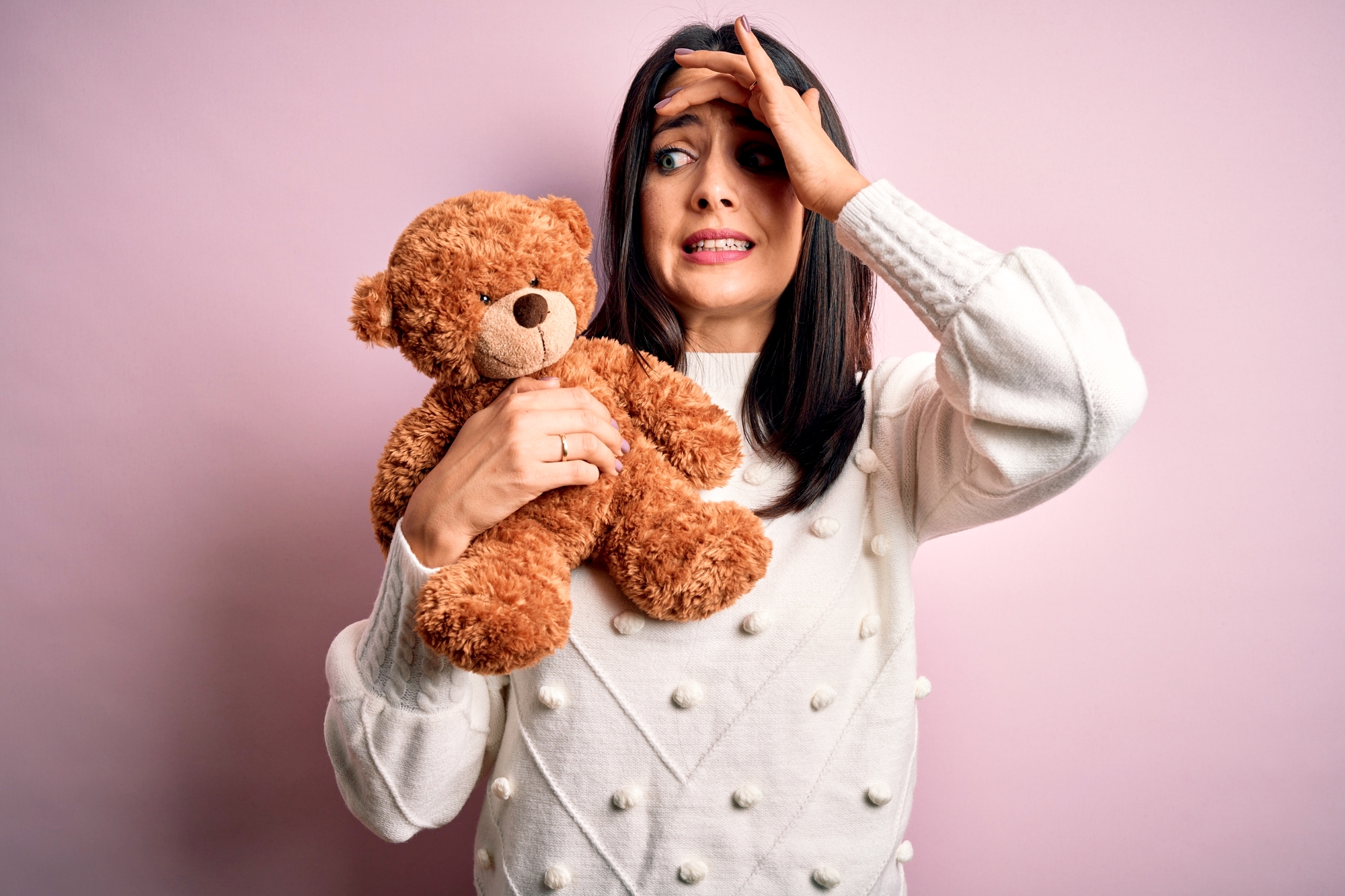 A woman with dark hair in a white sweater nervously holds a brown teddy bear. Her left hand is on her forehead, and her face shows an expression of worry or concern. She stands against a pink background.