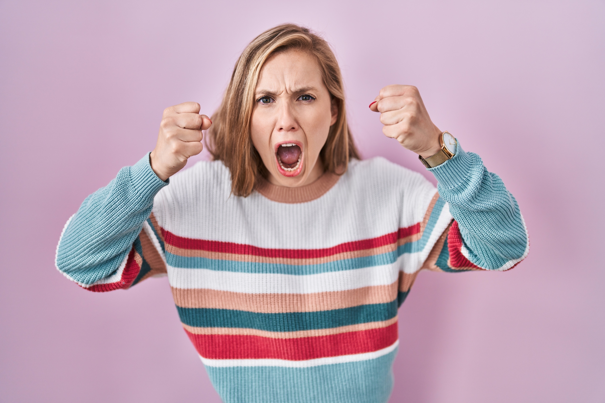 A woman with shoulder-length blonde hair is standing against a light pink background. She is wearing a colorful striped sweater and a watch on her wrist. She looks angry, with her mouth open and fists raised.