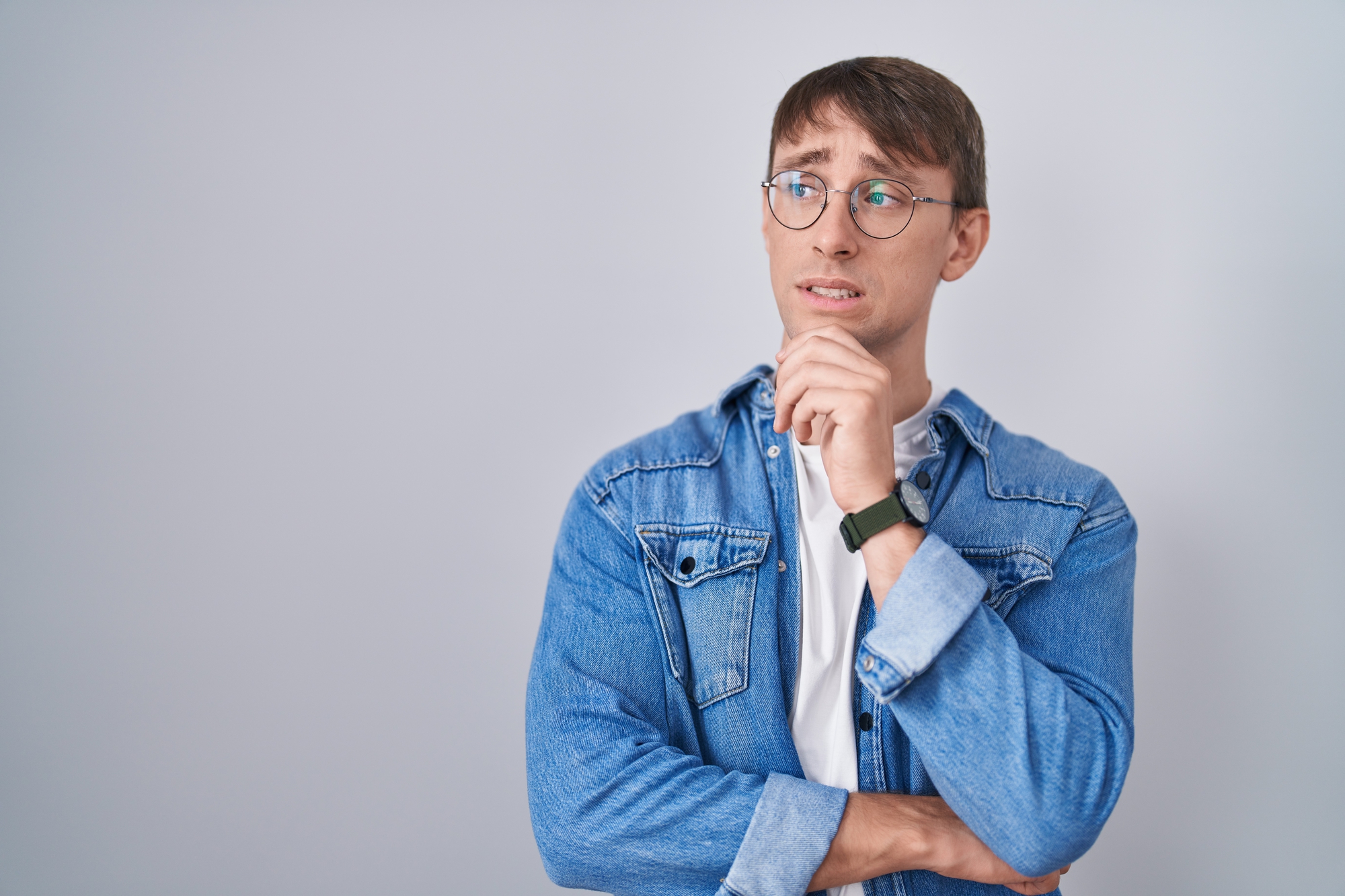 A man with short brown hair wearing round glasses, a denim jacket, and a white T-shirt stands against a plain light gray background. He has one hand resting on his chin and looks to the side with a thoughtful expression.