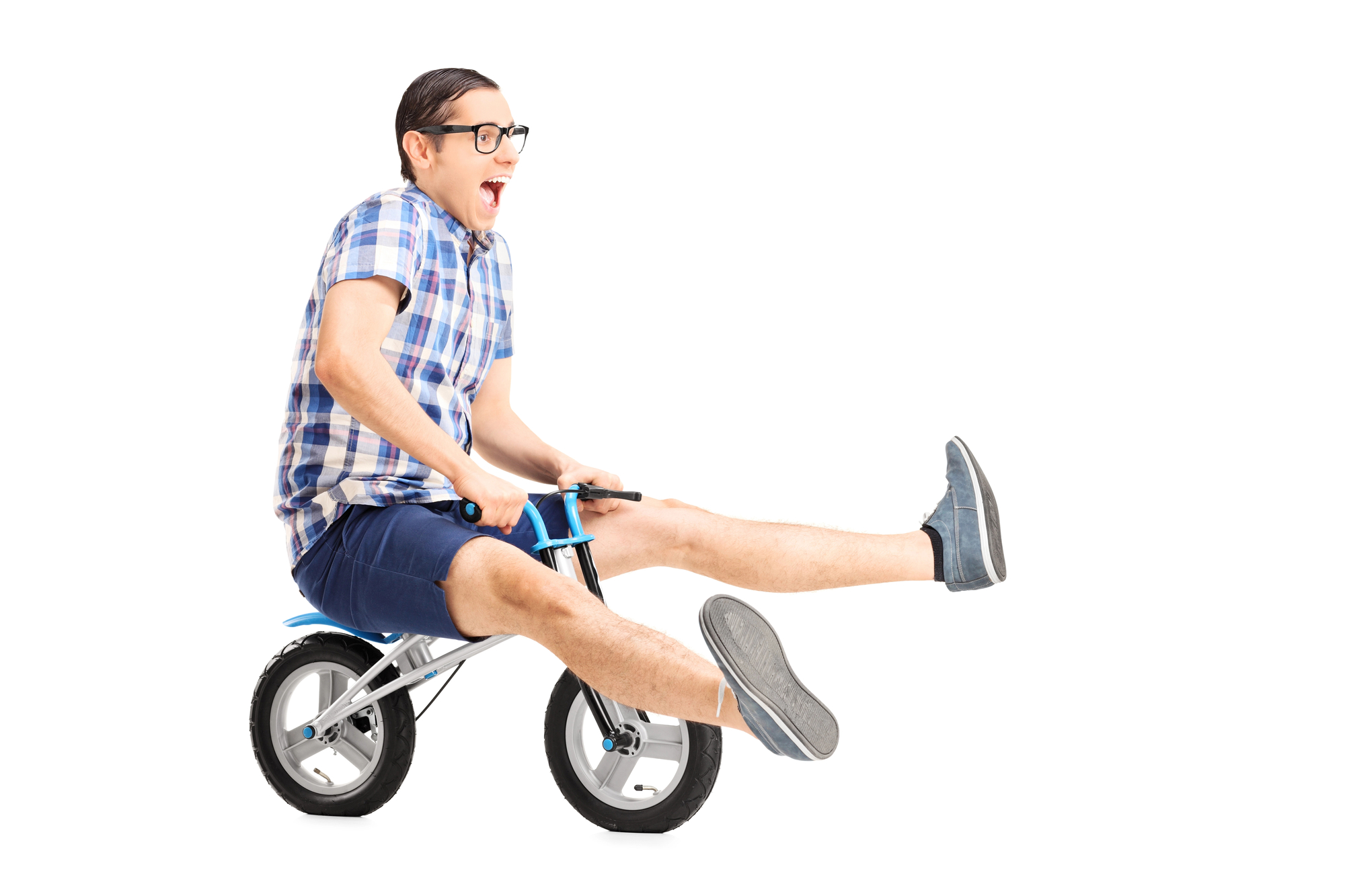 A joyful man in glasses and a plaid shirt rides a small children's bike. He has his legs extended forward with excitement and is wearing casual shorts and shoes. The background is plain white, highlighting his playful expression and the humor of the situation.
