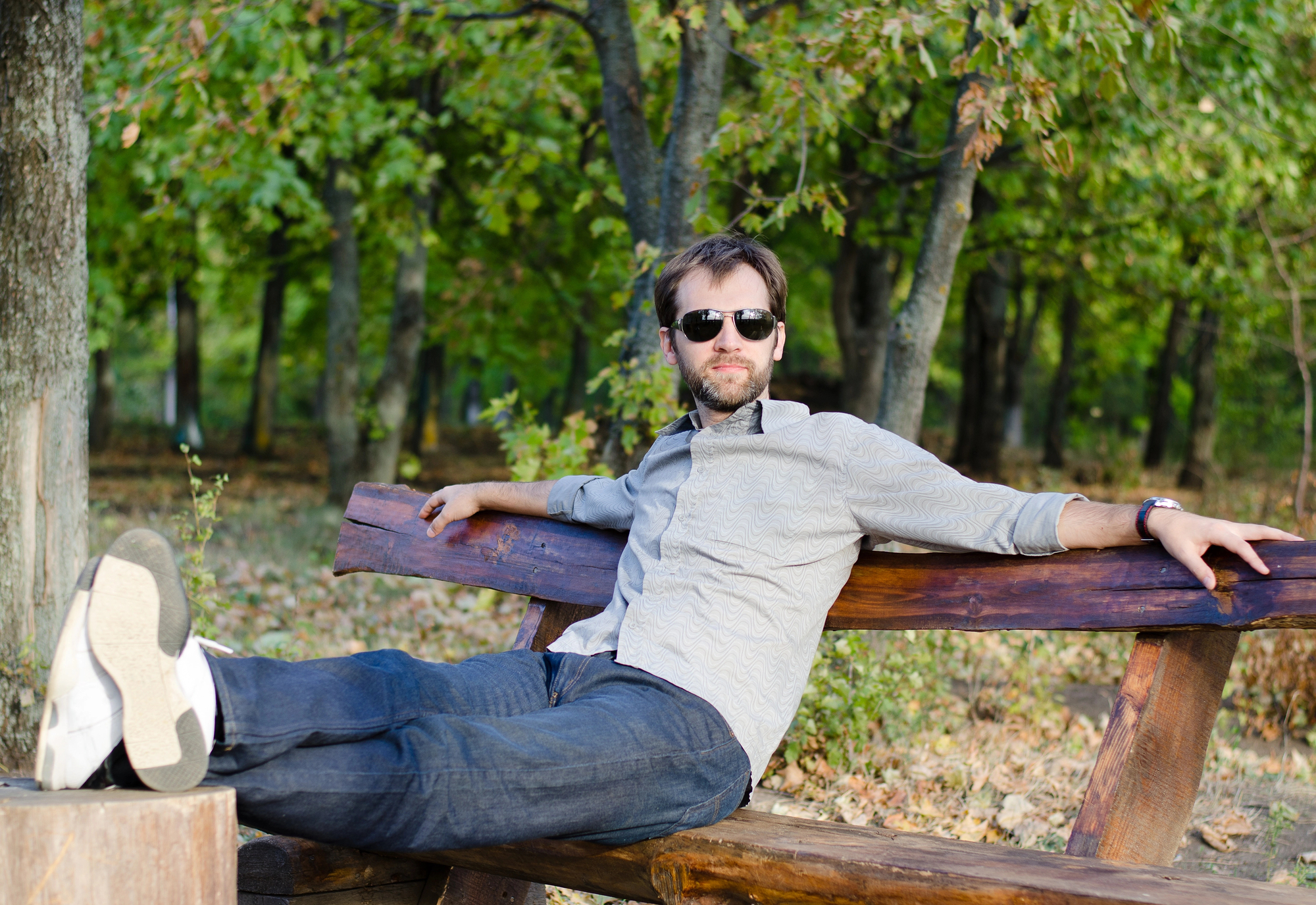 A man in sunglasses and casual clothing lies relaxed on a wooden bench in a park, with one leg resting on the bench and the other stretched out. The background features green trees, creating a serene, natural setting.