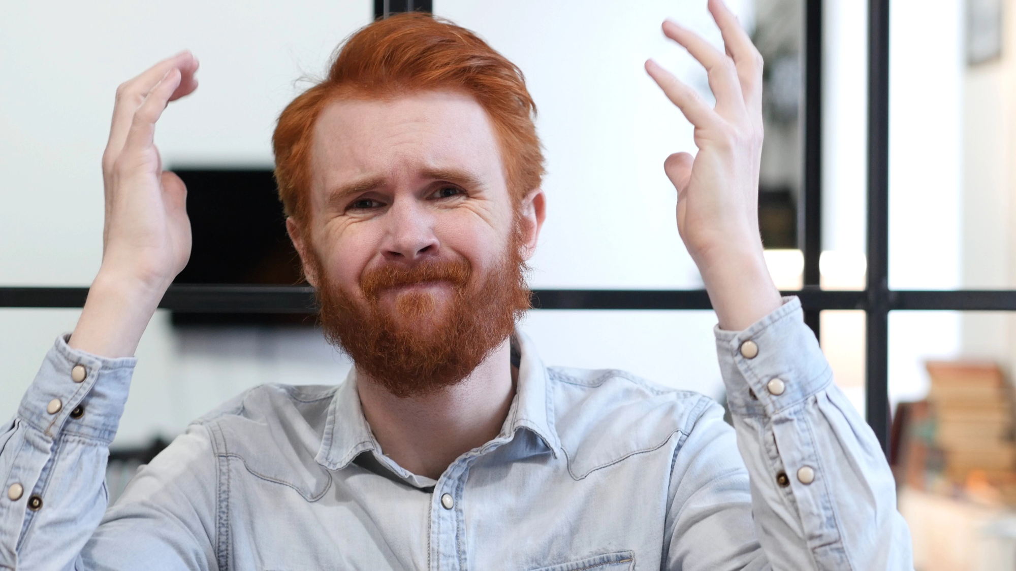 A person with red hair and a beard, wearing a light denim shirt, is sitting with a frustrated expression, raising both hands beside their head. The background shows a blurred indoor setting.