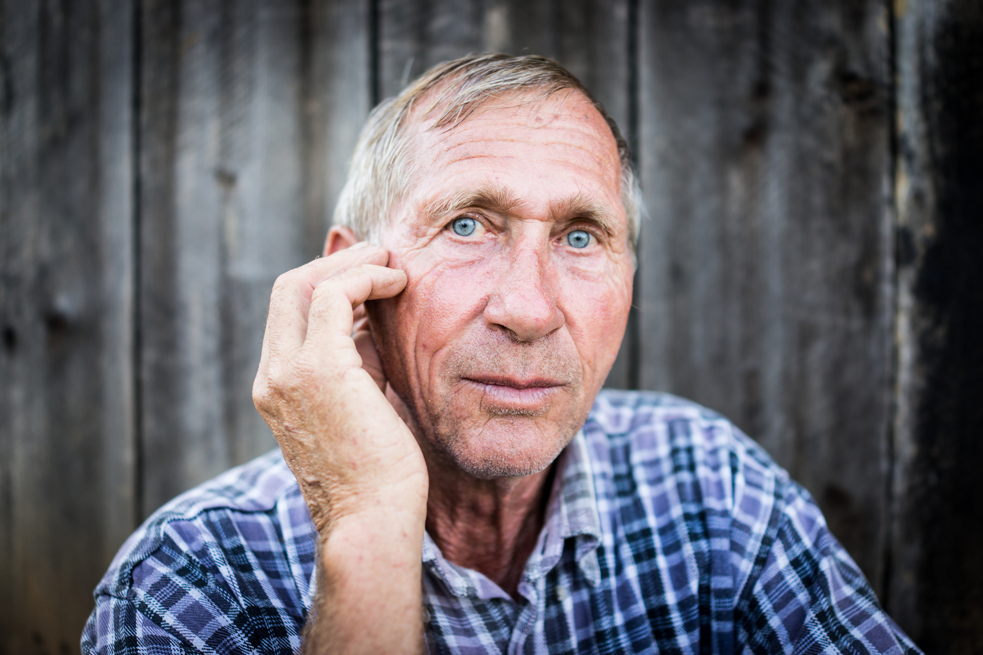 An elderly man with short gray hair and blue eyes, wearing a plaid shirt, looks directly at the camera. He holds his right hand to the side of his face near his eye. The background is a weathered wooden fence.