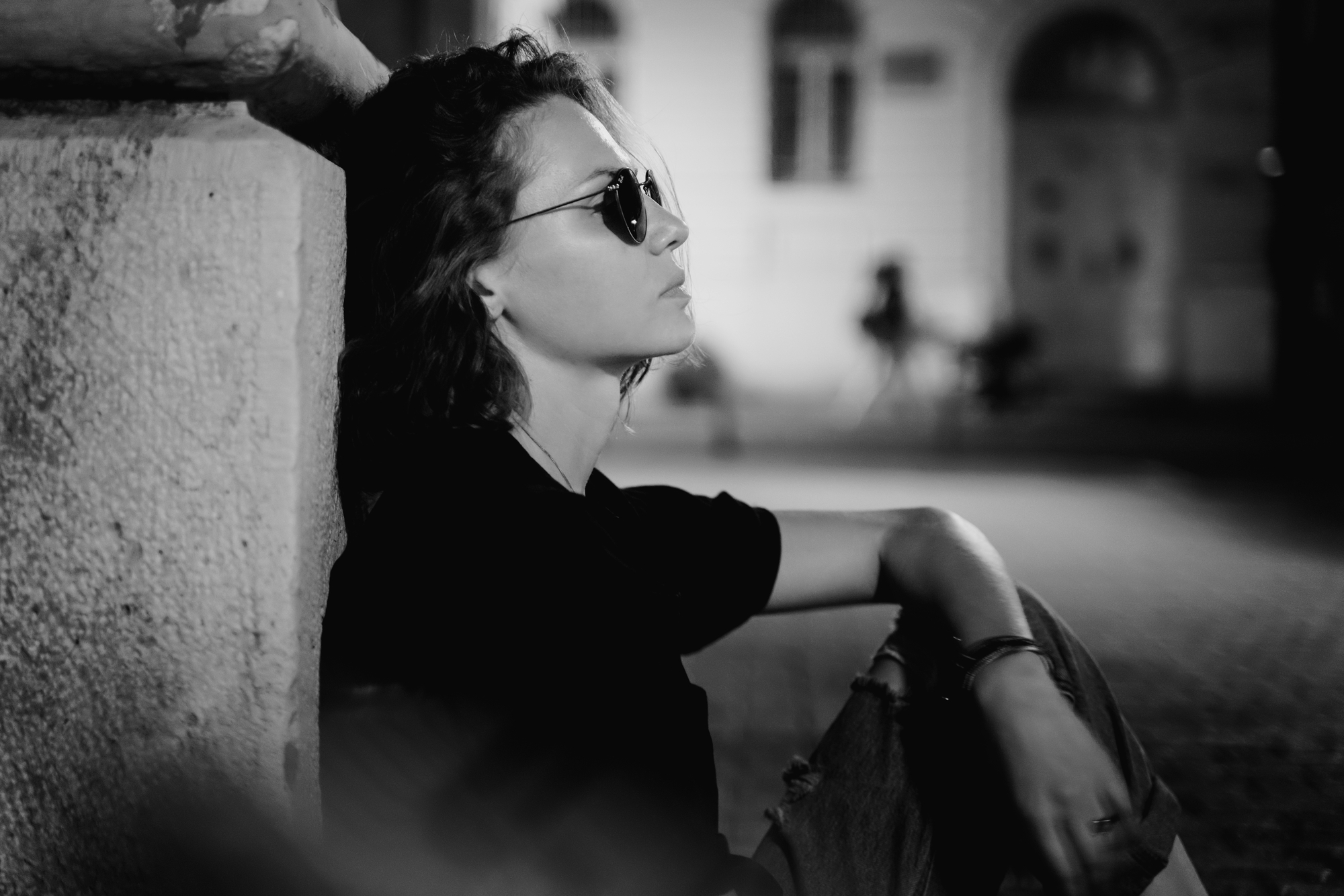 Black and white photo of a person with short hair sitting against a wall, looking thoughtful. They are wearing sunglasses, a dark shirt, and jeans. The background is blurred, suggesting a quiet urban setting at night.