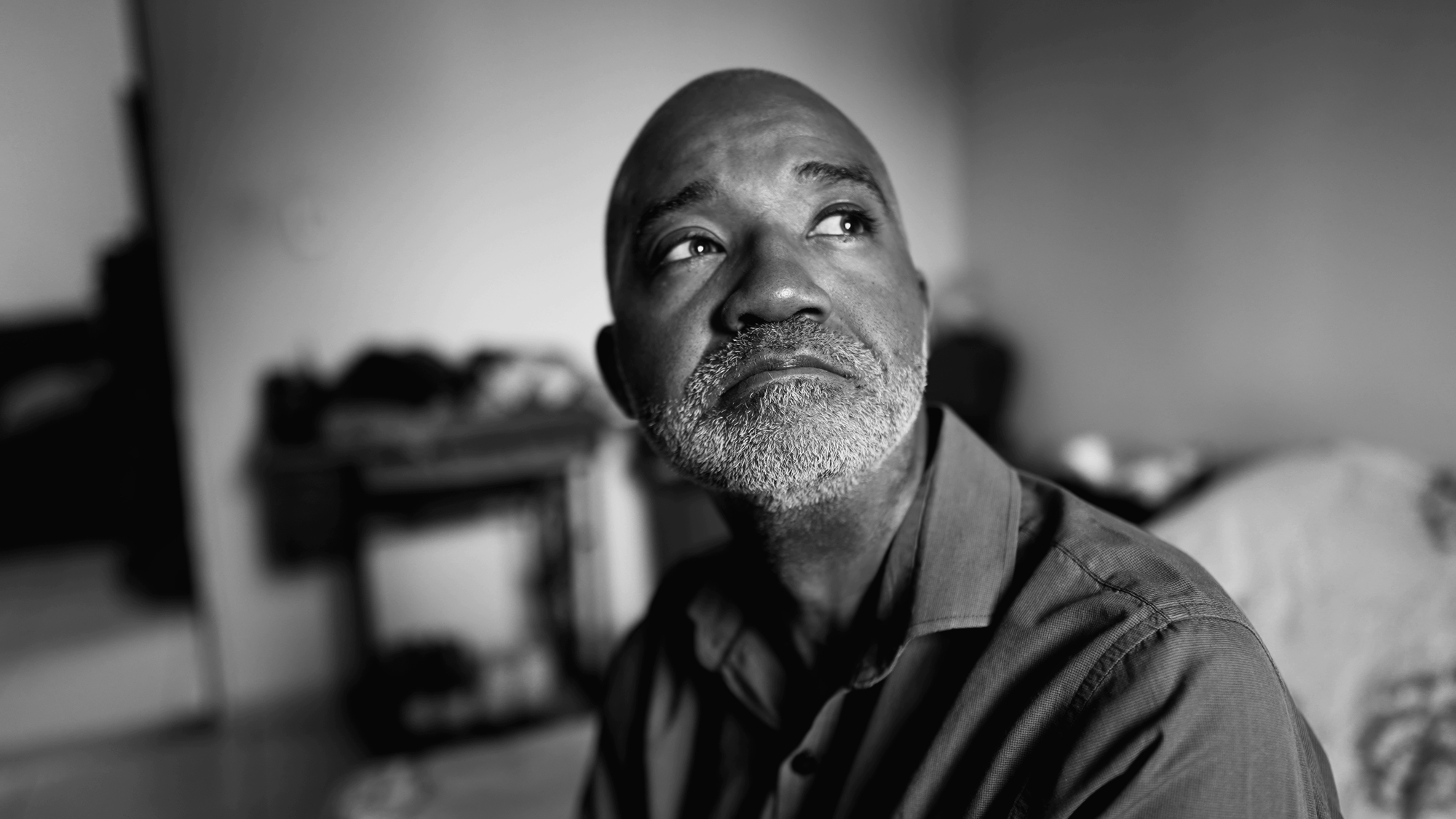 A grayscale close-up of a man with a bald head and gray beard. He wears a collared shirt and gazes thoughtfully upward to his right. The background is softly blurred, showing indistinct indoor surroundings like furniture and other objects.