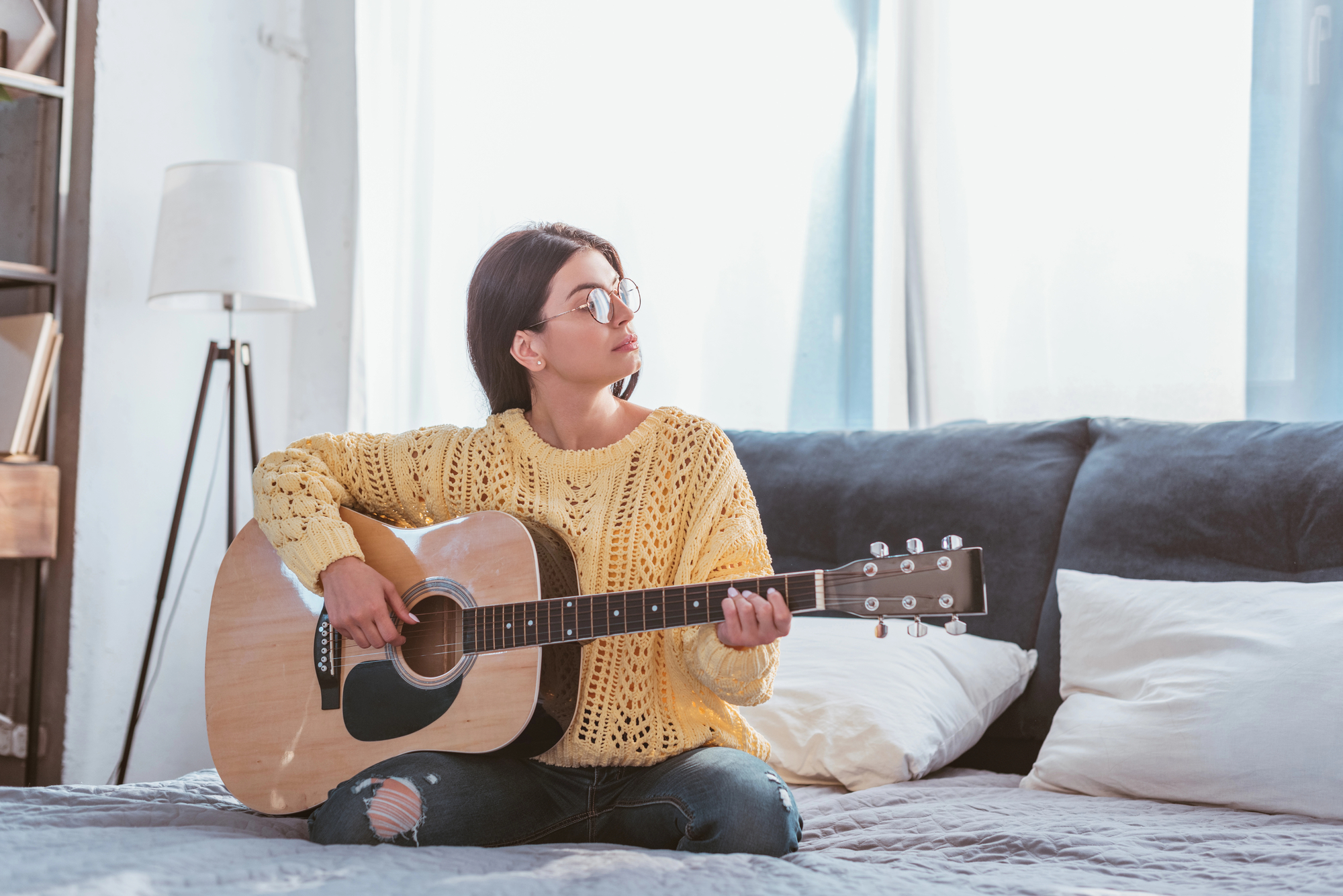 A person with glasses sits on a bed, wearing a yellow sweater and ripped jeans, playing an acoustic guitar. The room has a bright, natural light from a window and a neutral, cozy decor with pillows and a lamp in the background.