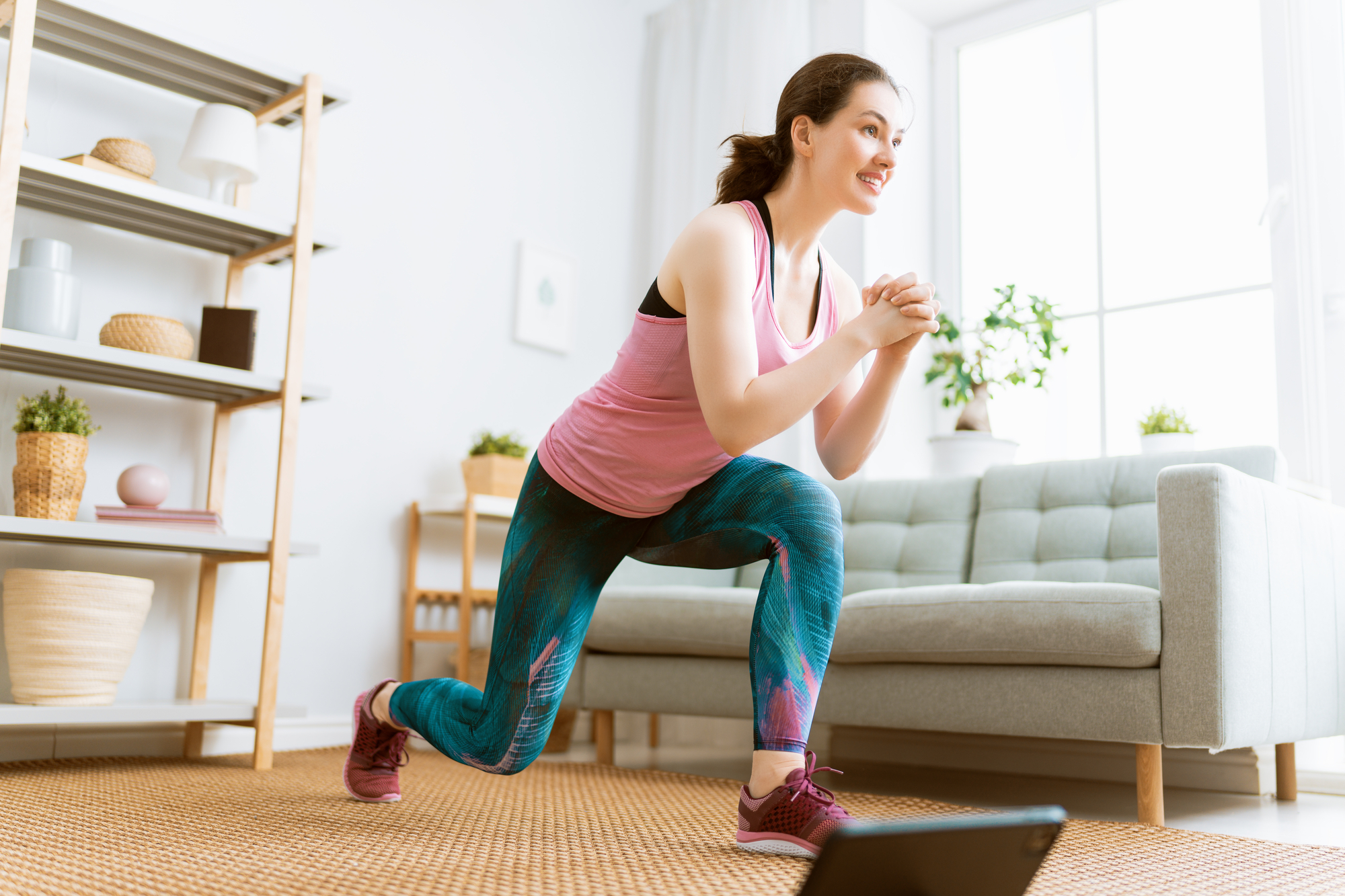 A woman in exercise clothes does lunges in a bright living room with a laptop in front of her. She is wearing a pink tank top and colorful leggings. The room has a sofa, wire shelf with decorative items, and a large window letting in natural light.