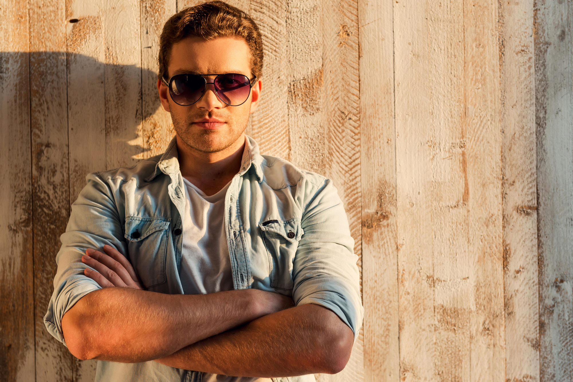 A man with short hair wearing dark aviator sunglasses and a light denim shirt stands with his arms crossed against a textured wooden wall, illuminated by warm sunlight from the left.