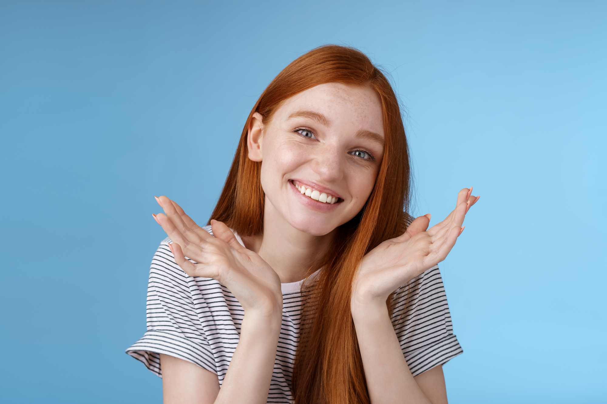 A smiling young woman with long red hair is wearing a striped t-shirt and holding her hands up near her face in a playful gesture. She is standing against a solid light blue background.