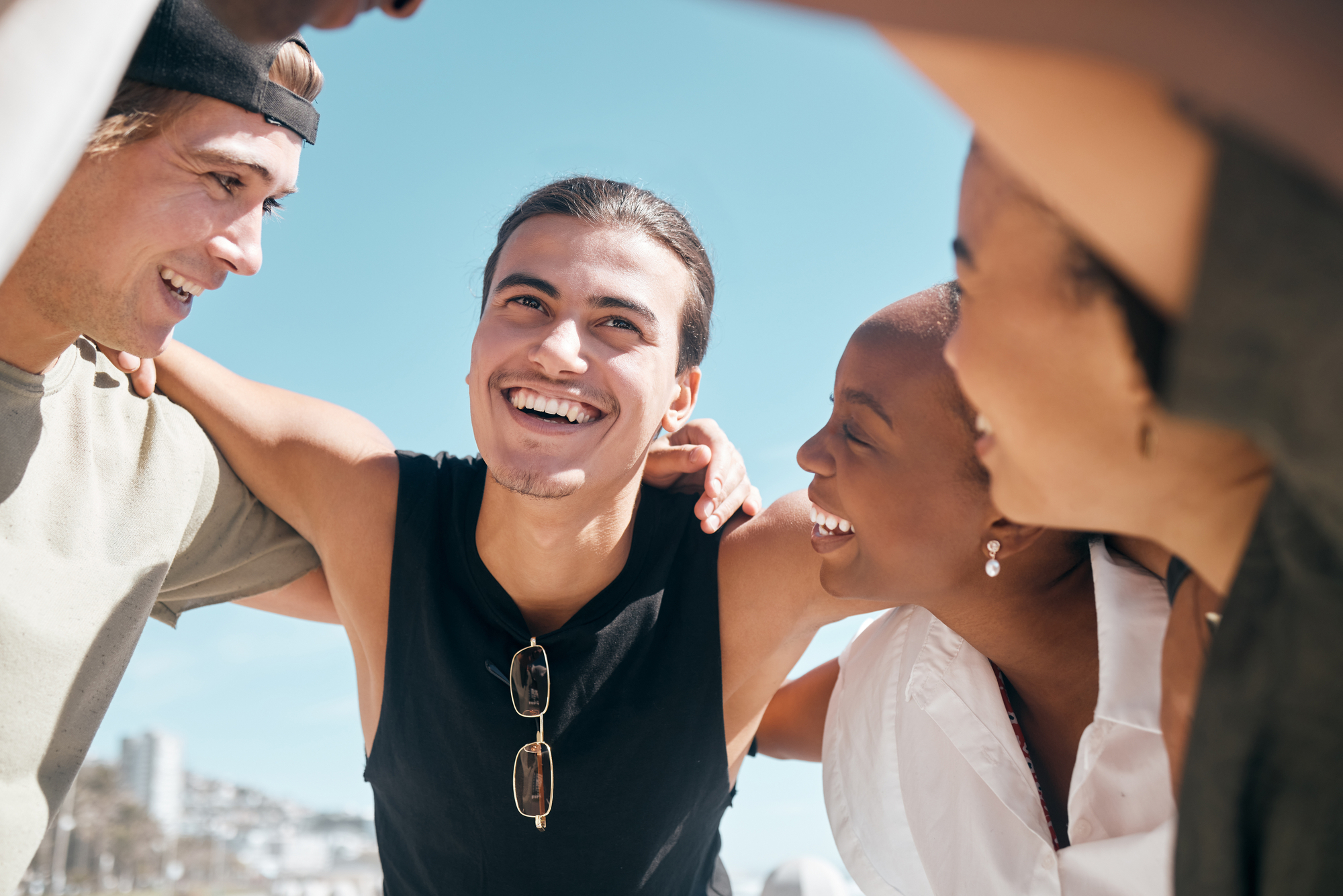 A group of four young friends stand closely together outdoors, smiling and laughing under a clear blue sky. They have their arms around each other’s shoulders, exuding happiness and camaraderie. The setting appears to be a sunny day at the beach.