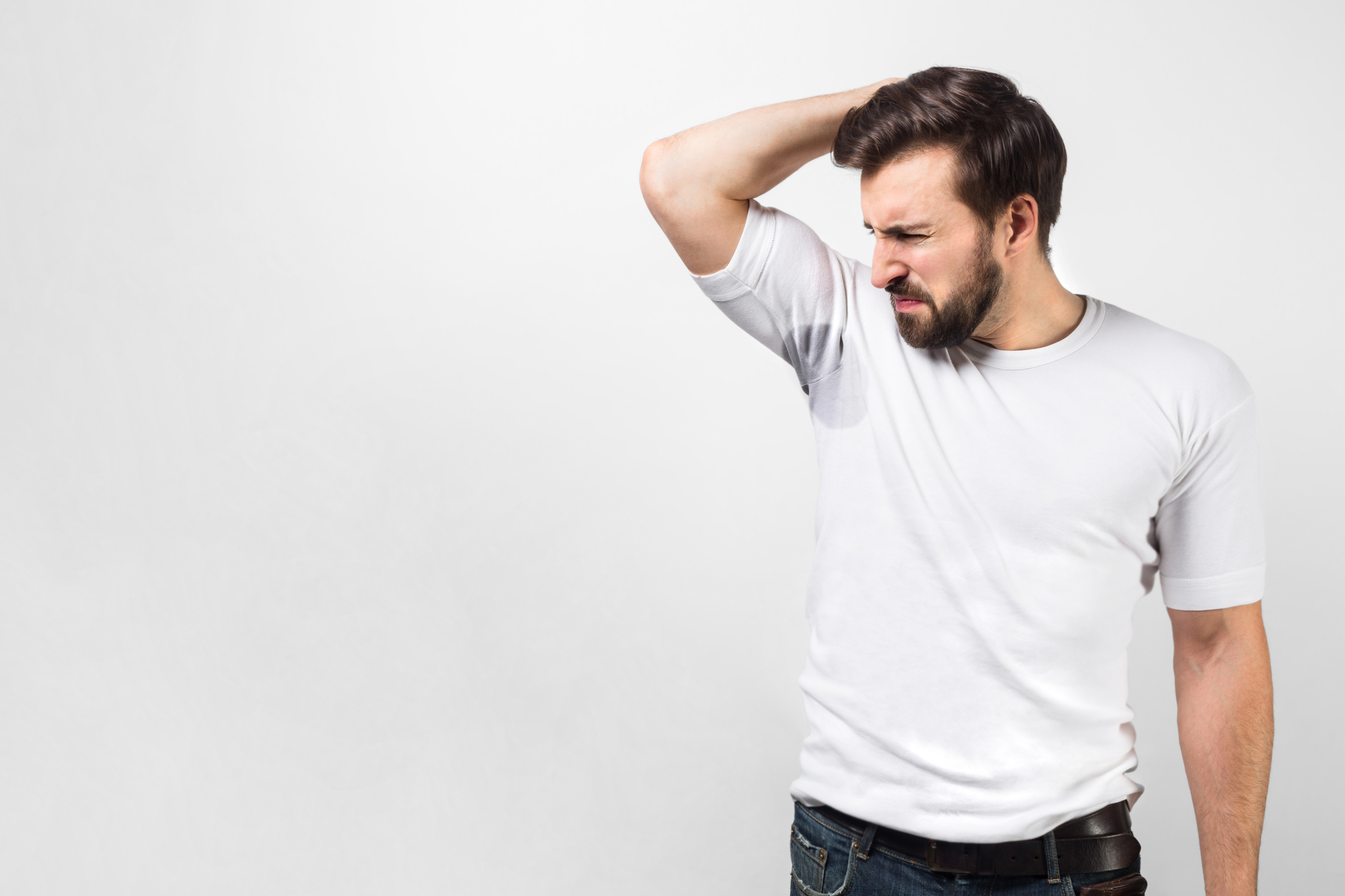 A man in a white t-shirt is standing against a plain white background. He has a pained expression on his face while raising his right arm to check his armpit, appearing to be concerned about sweat or body odor.