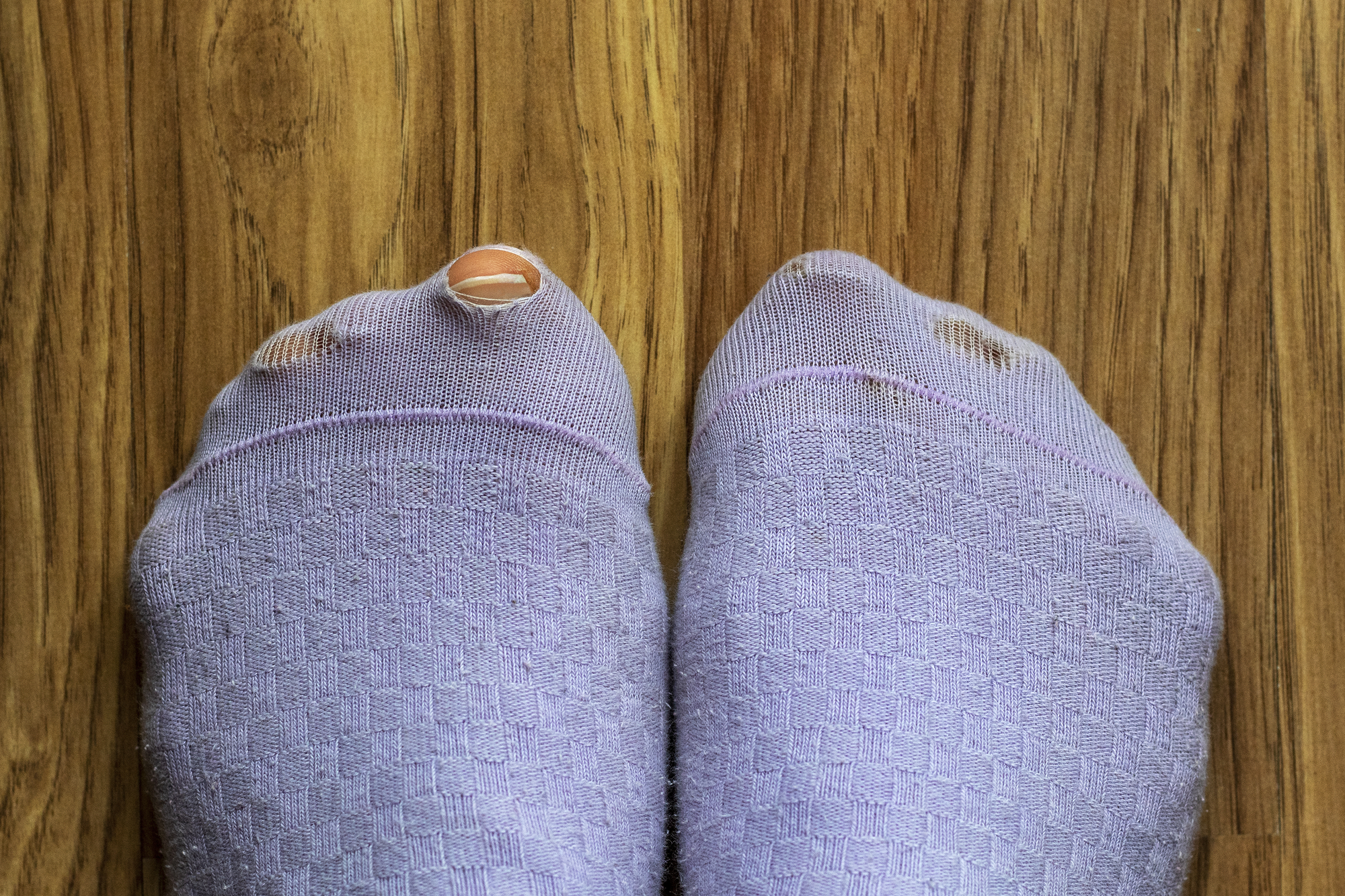 A pair of feet wearing worn-out, light purple socks with holes. The big toe on the left foot is poking through a hole, and the socks are against a wooden floor background.
