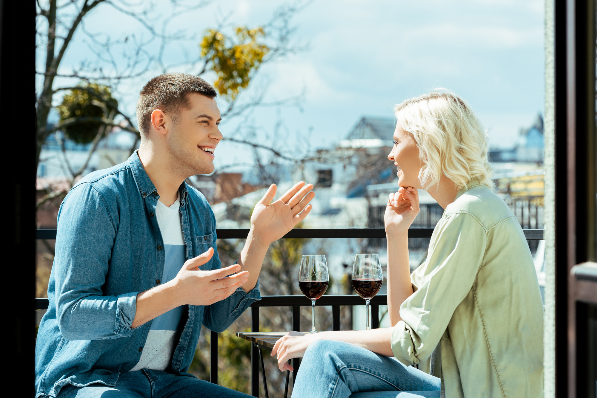 Two people sit on a balcony, engaged in a lively conversation. They are facing each other, smiling and gesturing with their hands. Two glasses of red wine are on a small table between them. The background shows trees and rooftops under a partly cloudy sky.
