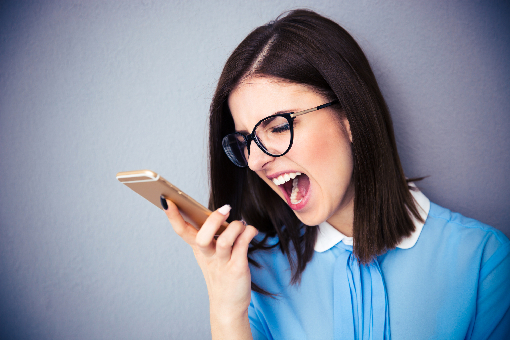 A woman with shoulder-length dark hair and glasses is leaning against a gray wall, holding a smartphone. She is wearing a light blue blouse and appears to be shouting into the phone, her mouth wide open in an expressive manner.