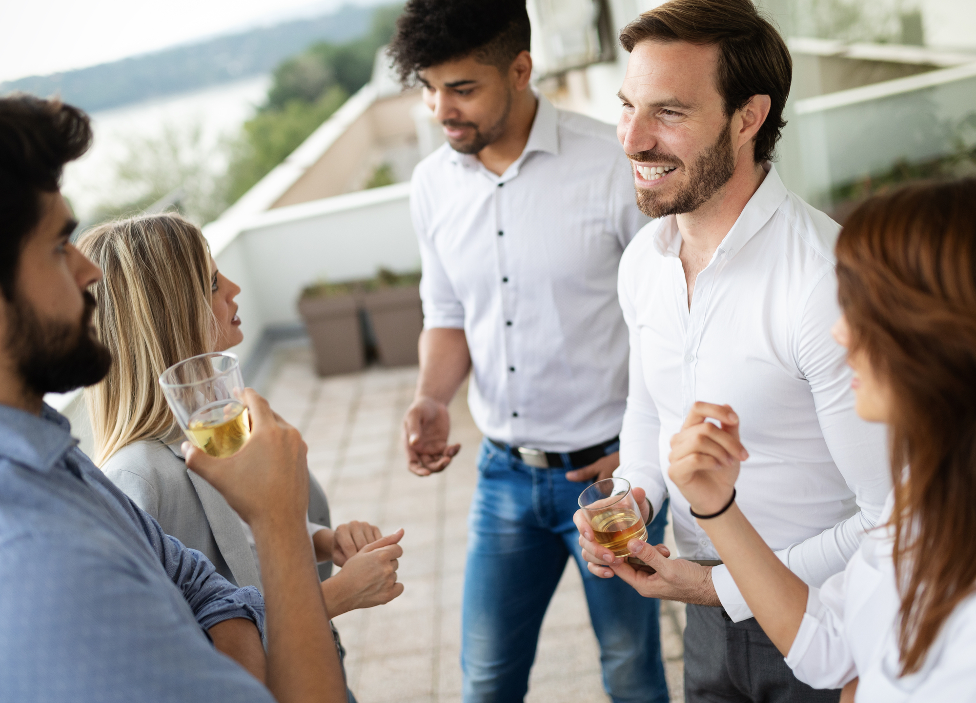 A group of five people casually talking and enjoying drinks on an outdoor terrace. They appear to be relaxed, with some smiling and holding glasses. The background includes a view of the landscape, featuring greenery and a body of water.