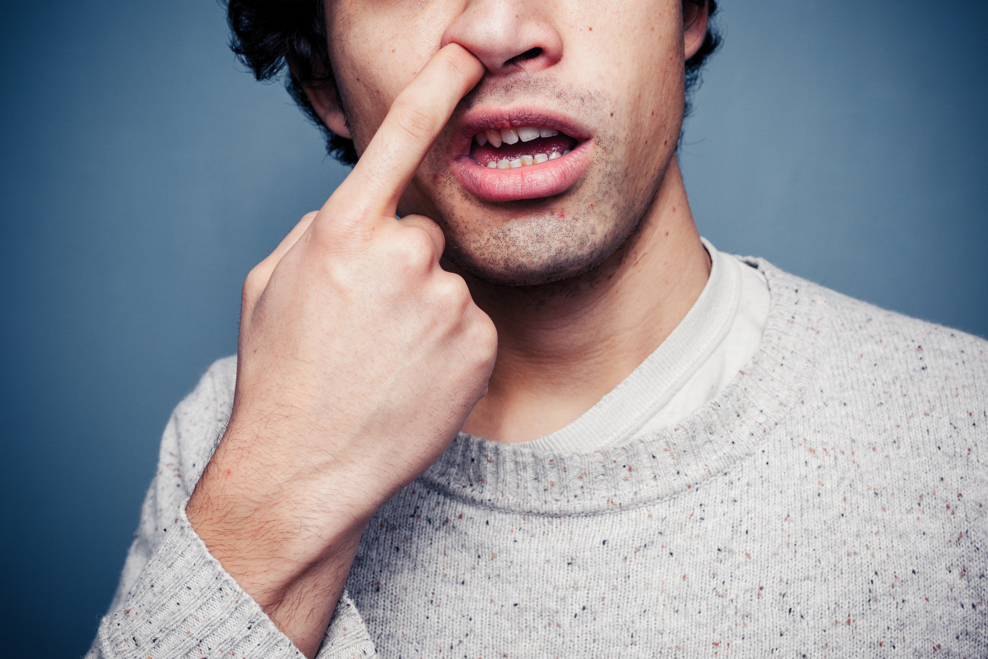 A person with dark curly hair and wearing a grey speckled sweater is picking their nose with their right index finger while partially opening their mouth. The background is a plain blue wall.