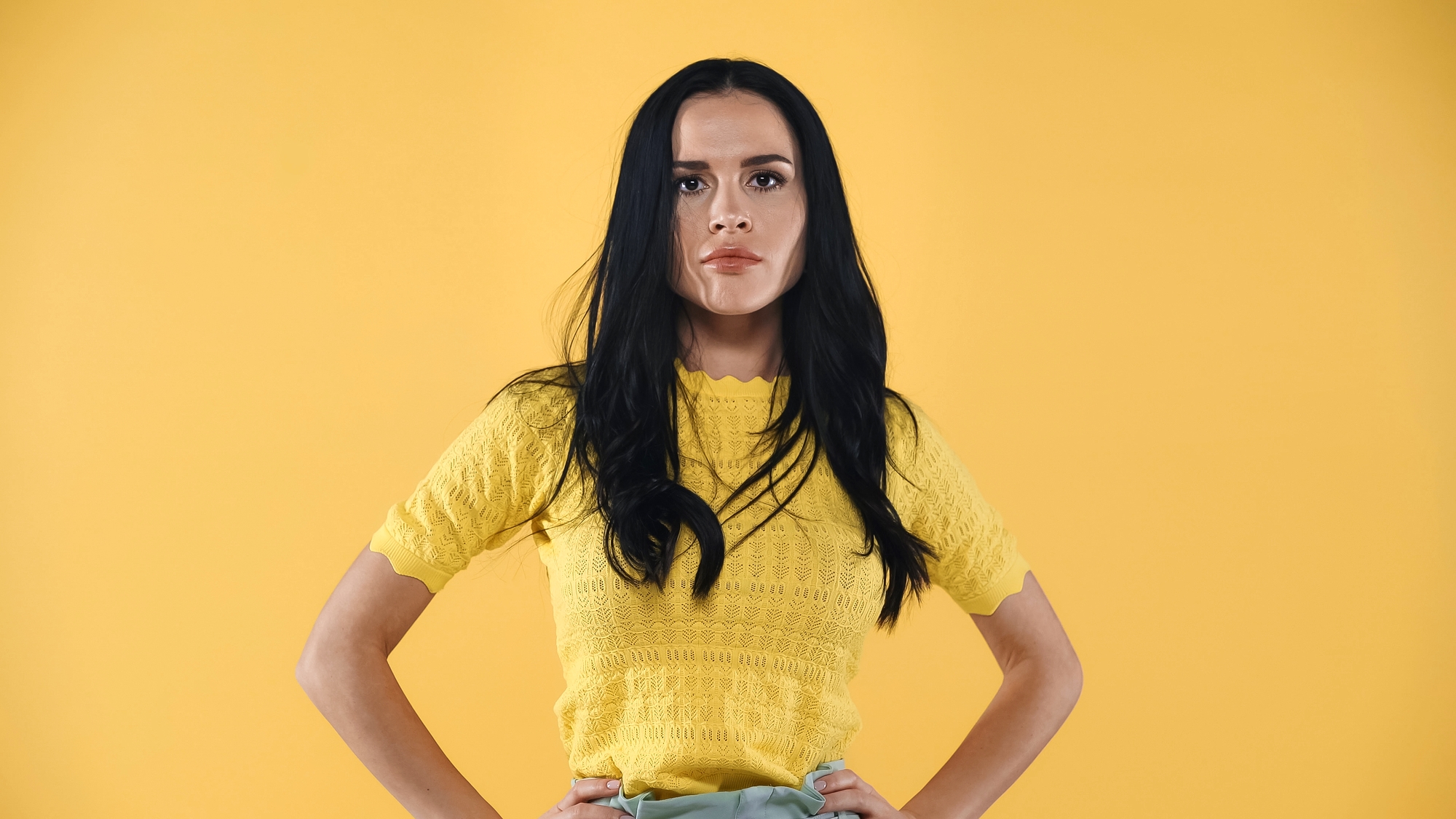 A person with long dark hair stands against a bright yellow background. They are wearing a yellow short-sleeved top and have their hands on their hips. Their expression is serious.