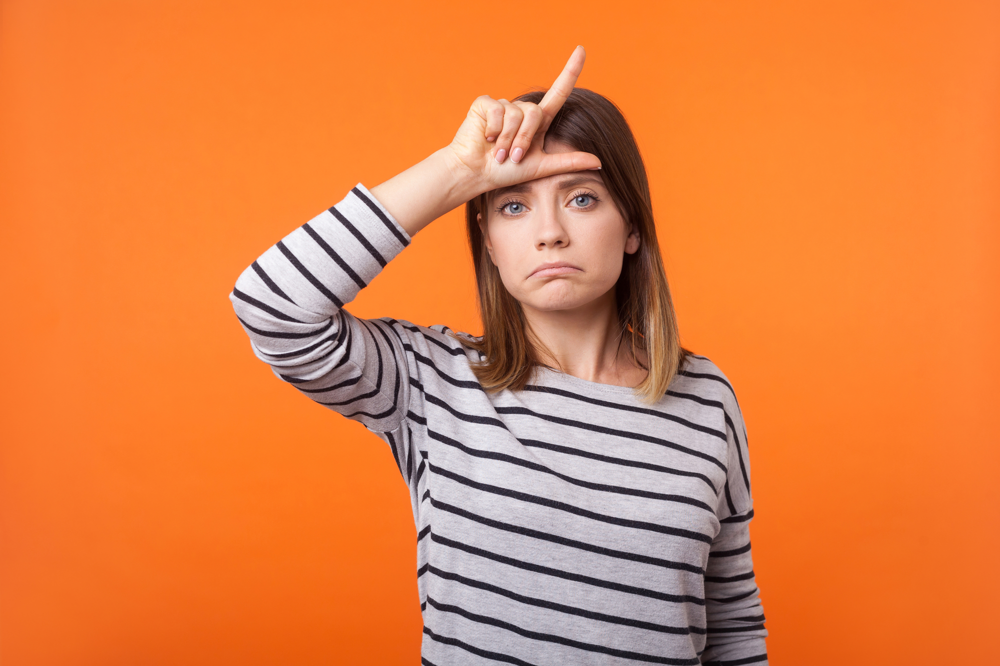 A woman with shoulder-length hair wearing a black and white striped shirt is standing against an orange background. She has her right hand raised to her forehead, forming an "L" shape with her fingers, and is making a disappointed or disapproving expression.