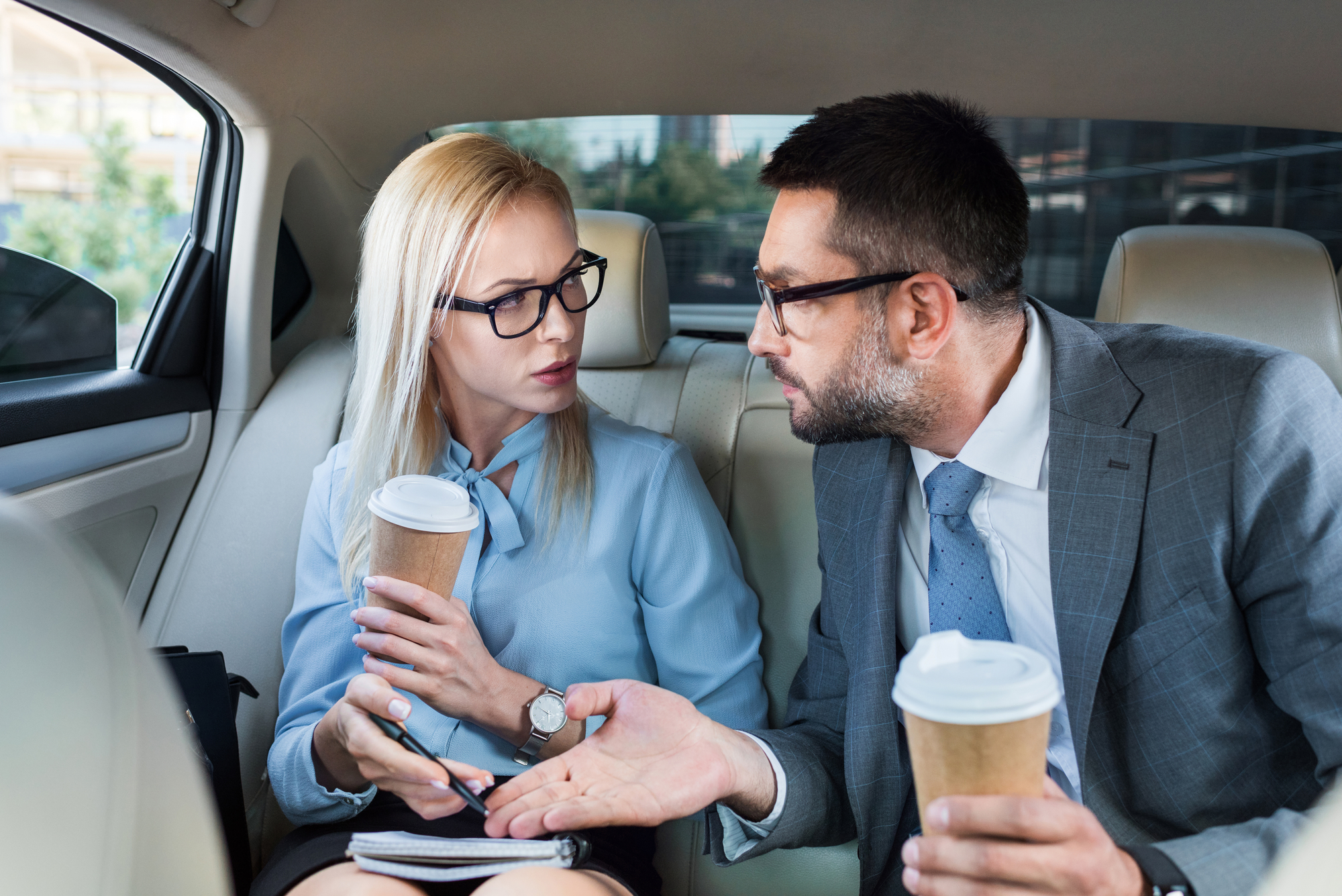 Two business professionals sit in the back seat of a car, both holding coffee cups. They are engaged in a serious conversation. The woman is writing in a notebook and the man is gesturing with his hand. They are both dressed in business attire and wearing glasses.