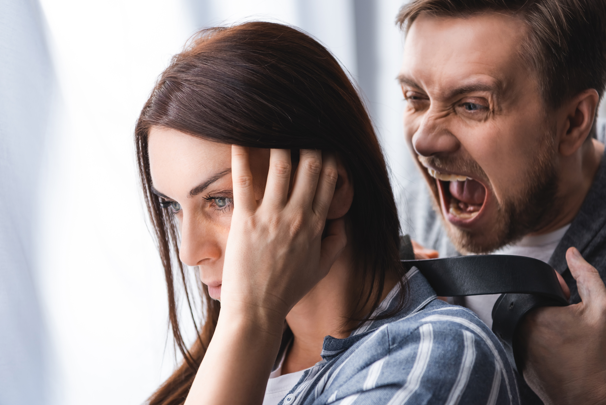 A close-up of a woman covering her ears with her hands, looking distressed and facing away. Behind her, a man is shouting aggressively, with his mouth wide open. Both are indoors with a neutral background. The scene conveys a sense of conflict and emotional distress.