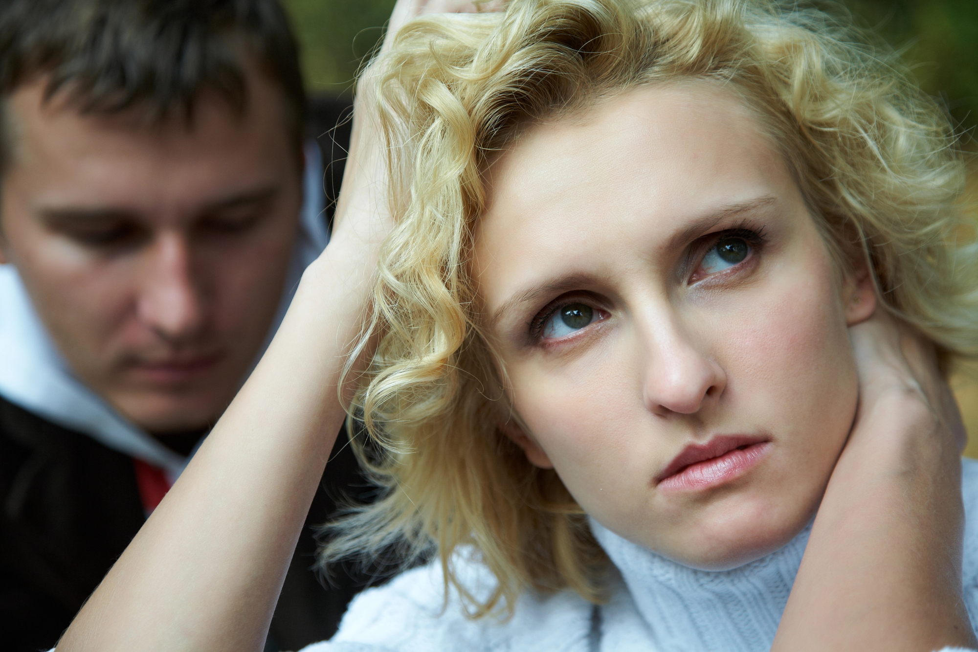 A blonde woman with curly hair looks pensive, gazing to the side with her hands touching her head. In the background, a man with short dark hair appears out of focus, his expression indistinct. The setting seems to be outdoors.