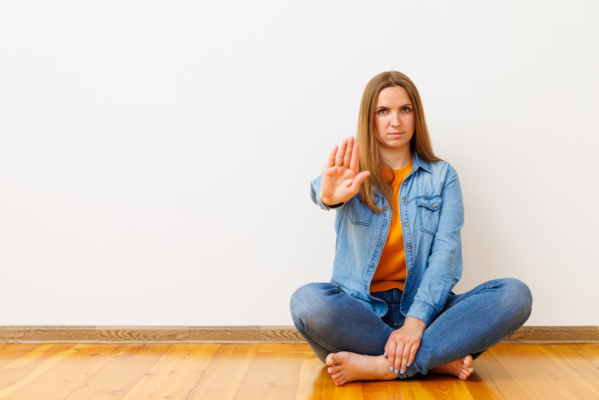 A woman with long hair sits cross-legged on a wooden floor against a plain white wall. She is wearing a denim jacket, jeans, and an orange top, with one hand raised in a stop gesture, her expression serious, conveying a clear message to halt or stop.