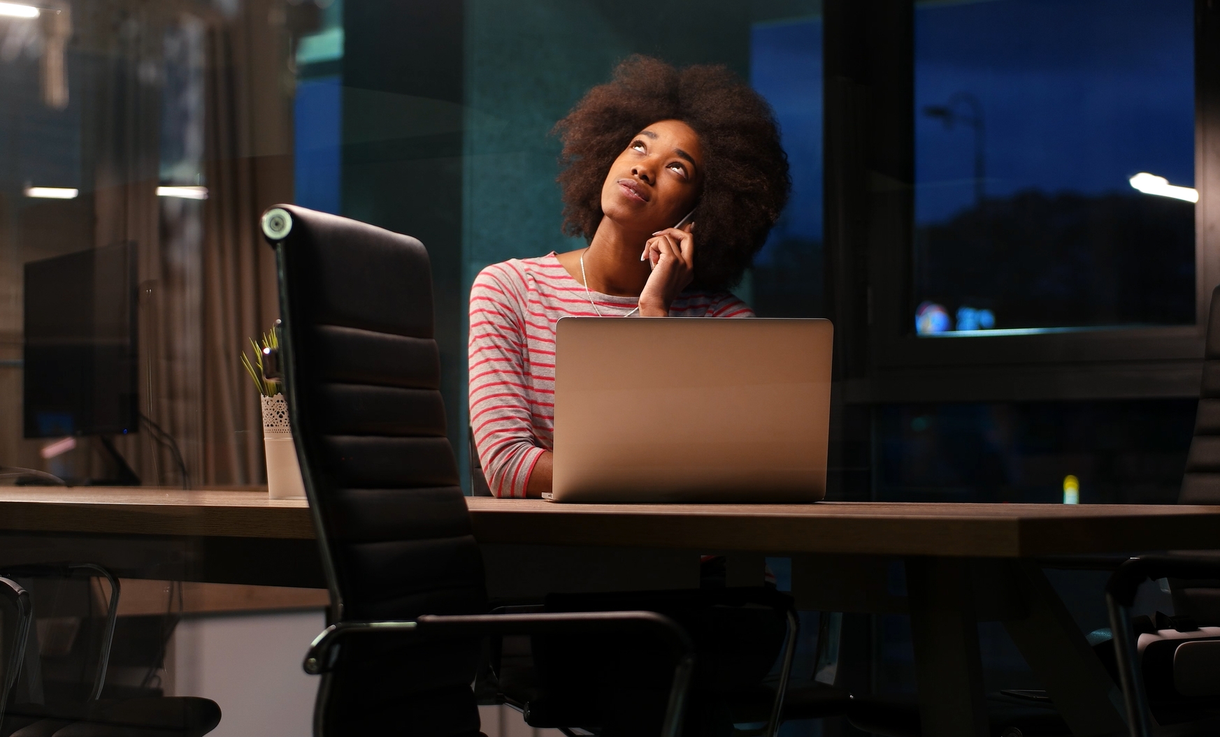 A person with curly hair, wearing a striped shirt, is looking up thoughtfully while sitting at a desk with an open laptop. The office environment around them is dimly lit, suggesting it's evening or night outside the window.