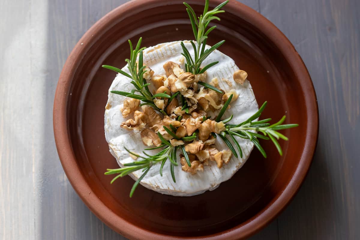 brie cheese is topped with rosemary sprigs and walnuts, and placed in a baking dish